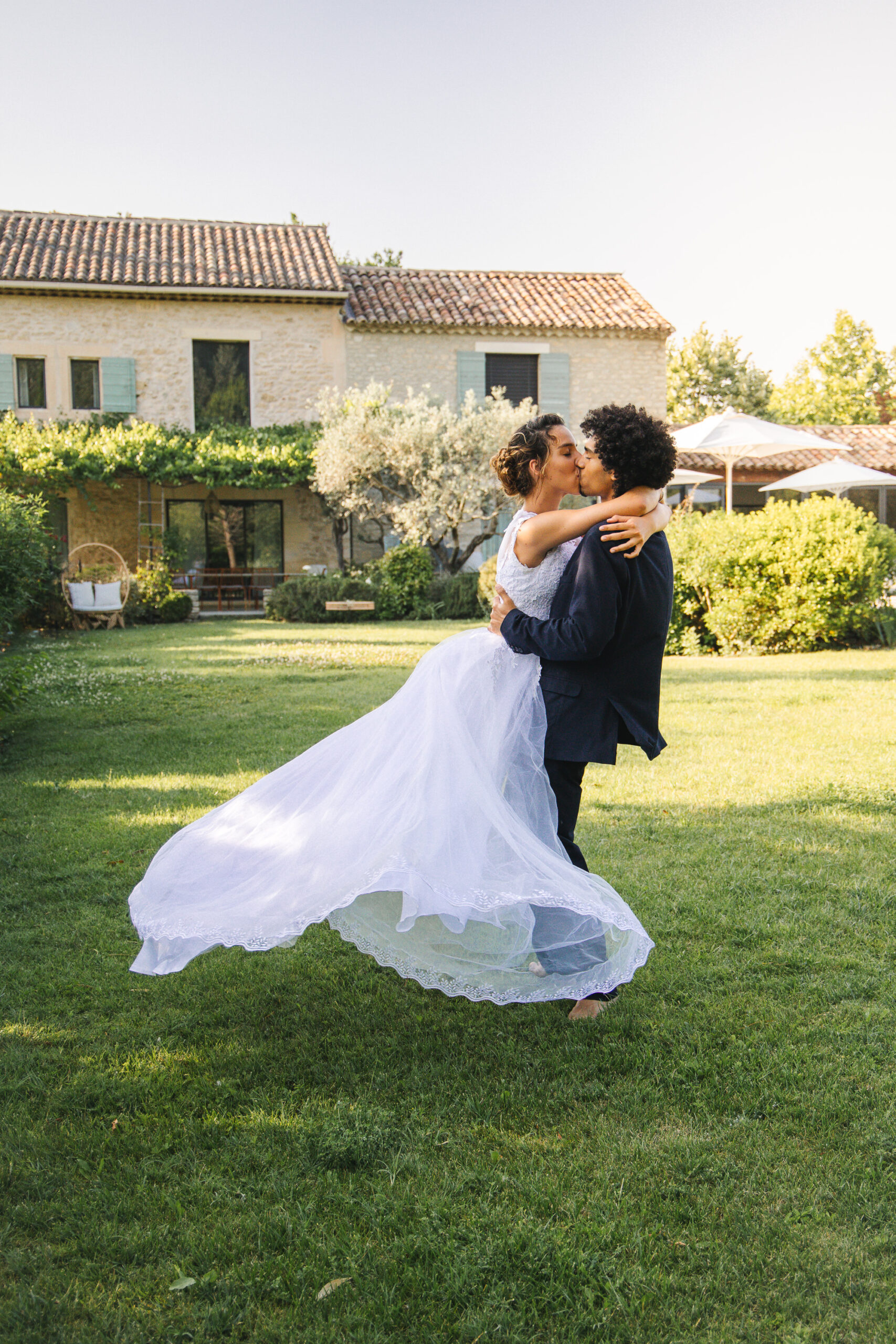 Mariée en robe blanche portée par son époux, robe en mouvement dans un jardin ensoleillé.