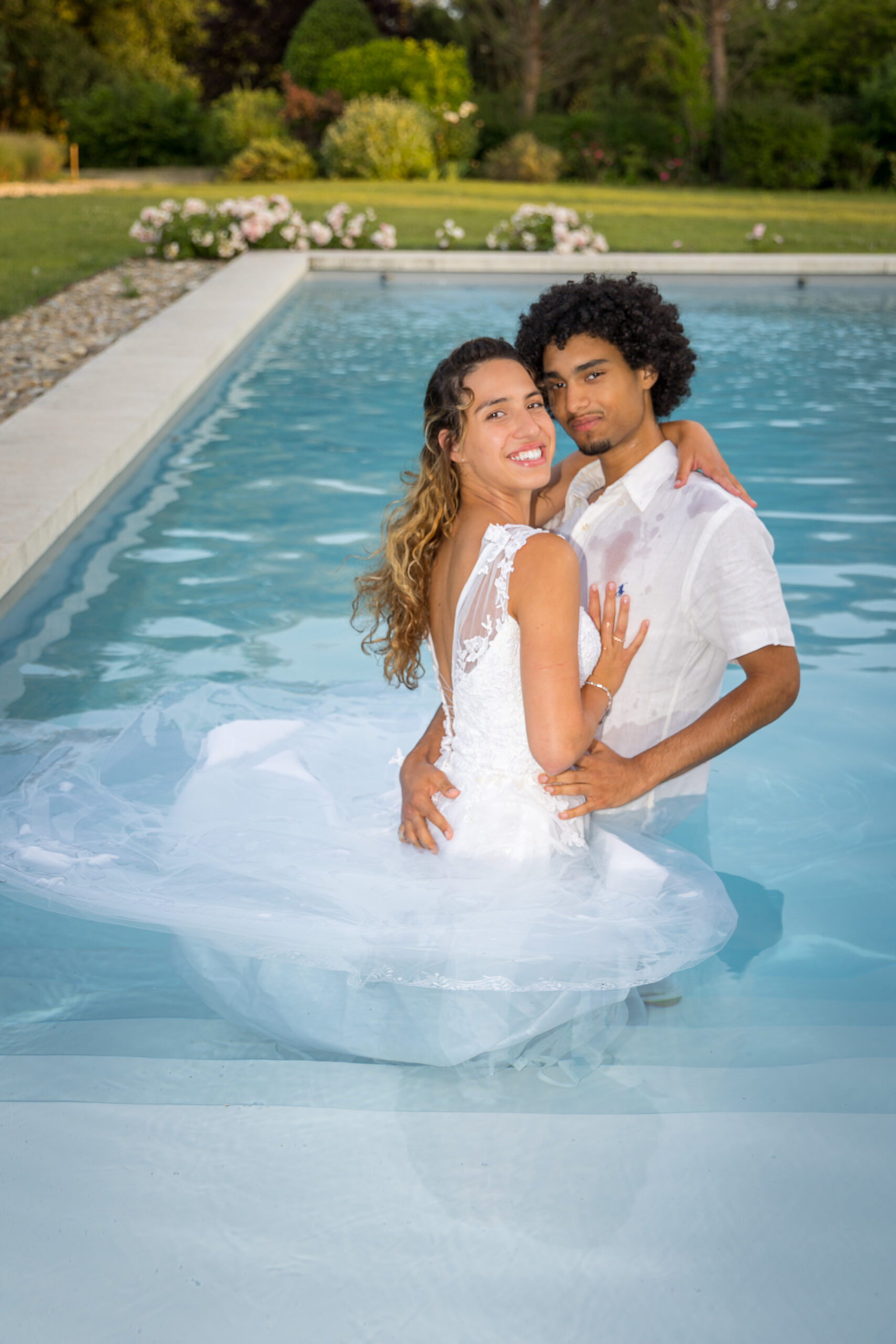 Un couple de mariés s’enlace tendrement dans une piscine, vêtus de blanc, le regard complice.