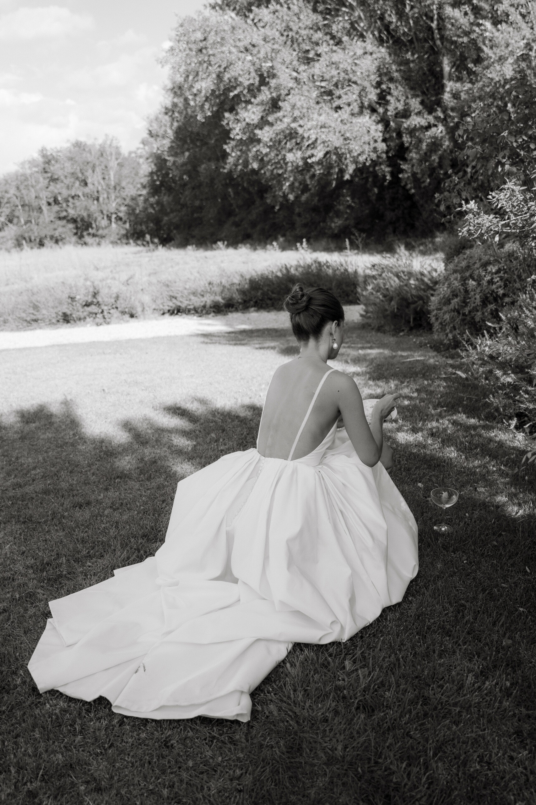 Mariée de dos assise au bord d’un cours d’eau en robe de mariée
