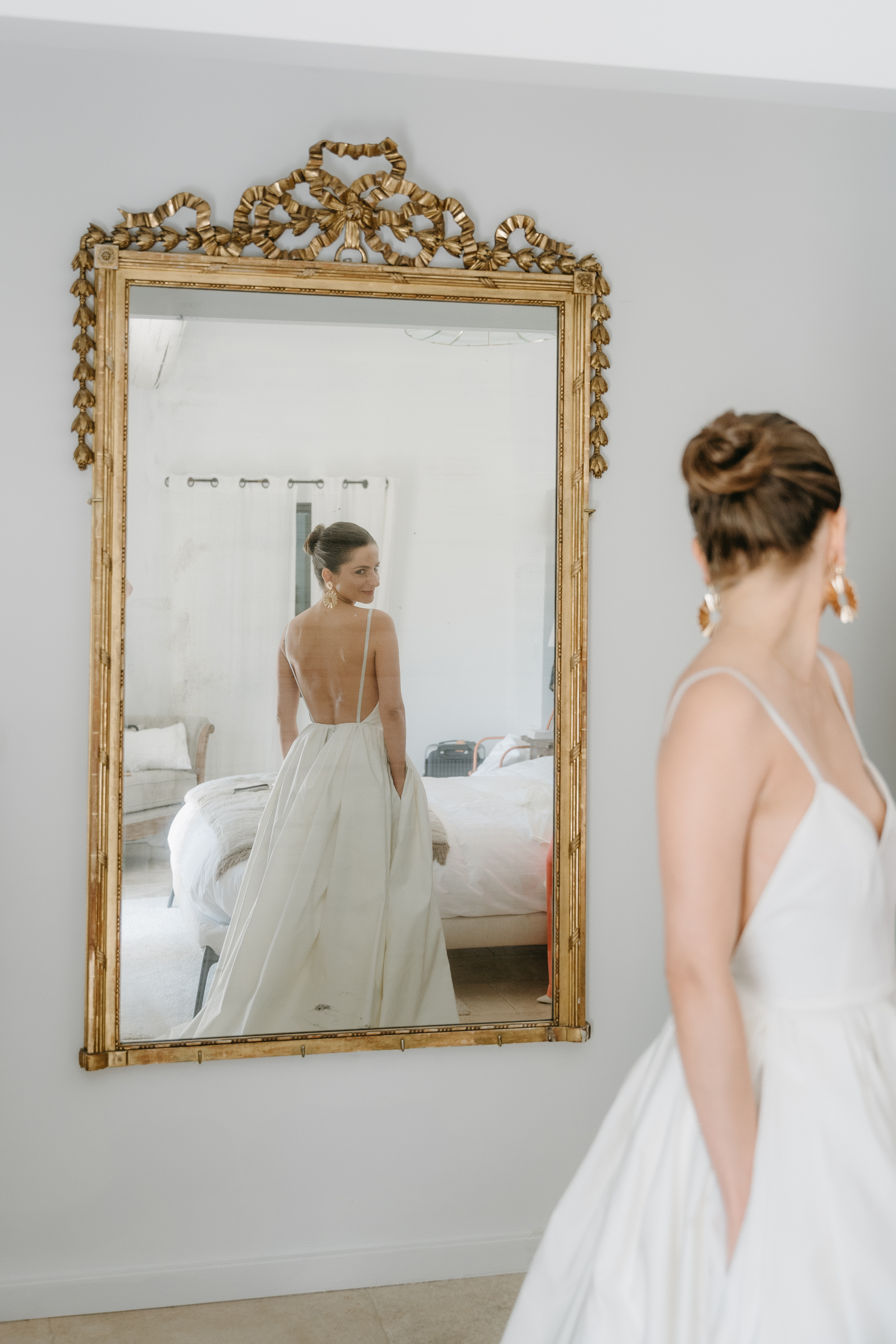 Mariée de dos face à un grand miroir doré, dans une robe élégante, se préparant pour la cérémonie.